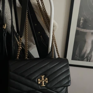 Svart quiltad axelväska med guldkedja - Snygg svart quiltad axelväska från Tory Burch med guldkedja och logotypdetalj på framsidan. Perfekt för att ge en lyxig touch till din outfit. Väskan har en elegant design med praktisk storlek för dina nödvändigheter. Knappt använd så jättefint skick!!😍