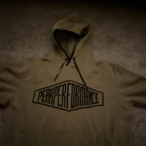 Olivgrön hoodie från Peak Performance - Säljer en olivgrön hoodie från Peak Performance med stor logga på bröstet. Den har en justerbar huva med svarta snören och en klassisk känguruficka framtill. Perfekt för en avslappnad stil.