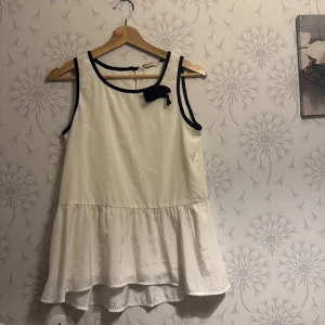 Vit ärmlös topp med svart rosett - Säljer en söt vit ärmlös topp med svart kant och en dekorativ rosett vid halsringningen. Toppen har en volangdetalj vid nederkanten som ger en fin siluett. Perfekt för en somrig look!