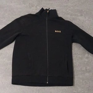 Svart jacka från Boss - Snygg svart jacka från Boss med dragkedja och broderad logga på bröstet. Jackan har långa ärmar och ribbade muddar vid ärmslut och nederkant. Perfekt för en stilren look.