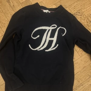 Svart tröja med logotyp från Tommy Hilfiger - Snygg svart tröja från Tommy Hilfiger med en stor vit logotyp på framsidan. Tröjan har långa ärmar och ribbade muddar vid ärmslut och nederkant. Perfekt för en stilren look.