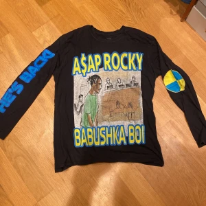 Svart långärmad tröja med A$AP Rocky-tryck - Cool svart långärmad tröja med färgglatt A$AP Rocky-tryck och texten 'Babushka Boi'. På ärmen finns ett blått och gult märke. Baksidan har text om en konsert i Stockholm. Perfekt för fans av A$AP Rocky!