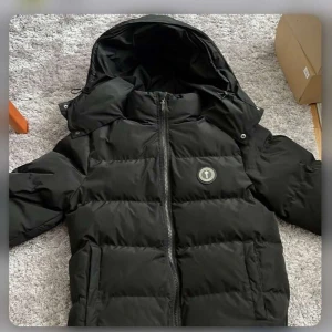 Svart pufferjacka från Trapstar - Snygg svart pufferjacka från Trapstar med huva och dragkedja. Jackan har en broderad logga på bröstet och text på ryggen. Perfekt för kyliga dagar. Knappt använd. Pris kan diskuteras vid snabb afärer