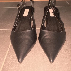 Svarta pumps från Nelly - Eleganta svarta pumps från Nelly med spetsig tå och justerbar rem runt hälen. Perfekta för en stilren look.