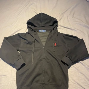 Svart hoodie från Polo Ralph Lauren - PRISET ÄR FAST!! Säljer en stilren svart hoodie från Polo Ralph Lauren med dragkedja och det klassiska röda logotypbroderiet på bröstet. Perfekt för en avslappnad look med ribbade muddar och justerbar huva.