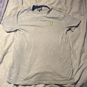 Grå t-shirt från Lyle & Scott Junior - Säljer en stilren grå t-shirt från Lyle & Scott Junior. Den har en liten gul broderad logga på bröstet och är kortärmad. Perfekt för en avslappnad look.