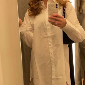Vit lång skjorta - Säljer en stilren vit långärmad skjorta med knappar framtill. Perfekt för en avslappnad men ändå elegant look. Säljer då den aldrig kommit till användning  