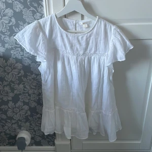 Vit blus med volangdetaljer - Charmig vit blus med korta ärmar och volangdetaljer. Blusen har en rund halsringning och knäppning bak. Perfekt för en somrig look.