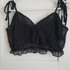 Svart prickig topp från NA-KD - Snygg svart topp från NA-KD med prickigt mönster och volangdetaljer. Toppen har knytbara axelband och en croppad passform. Perfekt för en trendig look!