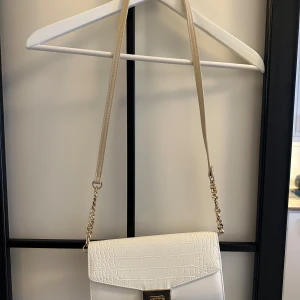 Vit axelväska från Anna Field - Snygg vit axelväska från Anna Field med krokodilpräglat mönster och guldfärgade kedjedetaljer. Perfekt för att ge en elegant touch till din outfit. Väskan har en praktisk storlek och en justerbar axelrem.