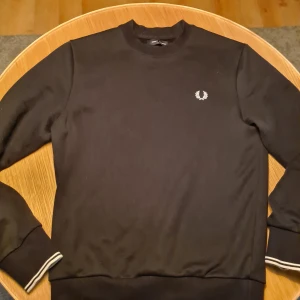 FRED PERRY CREW NECK - Säljer en stilren svart tröja från Fred Perry med deras ikoniska logga på bröstet. Nypris 1500kr. Öppen för prisförslag!