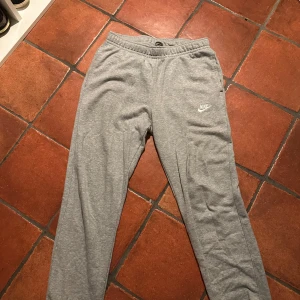 Grå mjukisbyxor från Nike - Säljer ett par grå mjukisbyxor från Nike i storlek M. Byxorna har en elastisk midja med snörning och en liten Nike-logga på benet. Perfekta för en avslappnad stil.