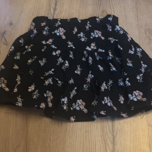 Svart blommig kjol från 157 New - Söt svart kortkjol med blommigt mönster från 157 New. Perfekt för en avslappnad stil med en elastisk midja för bekväm passform. Blommorna är i mjuka pastellfärger som ger en romantisk touch.