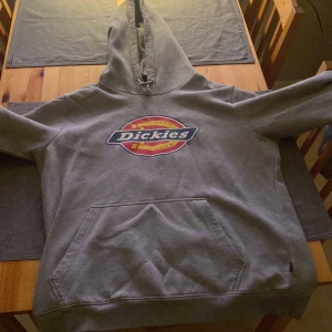 Grå hoodie från Dickies - Säljer en grå hoodie från Dickies med deras ikoniska logga i rött, gult och blått på bröstet. Tröjan har en stor ficka framtill och en justerbar huva. Perfekt för en avslappnad stil.