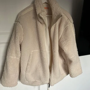 Beige teddyjacka från New Yorker  - Mysig beige teddyjacka från newyorker med dragkedja framtill. Jackan har en hög krage och långa ärmar, perfekt för kyliga dagar. Den har en avslappnad passform och två praktiska fickor på framsidan. Något konstigt med dragkedjan
