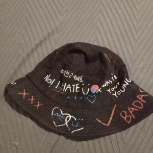 Svart bucket hat med text - Cool svart bucket hat med olika texter och symboler i vitt och rött. Perfekt för en edgy stil och attityd. Passar bra till avslappnade outfits.