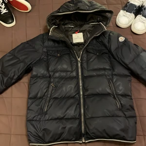 Mörkblå dunjacka från Moncler - Just nu så säljer jag min otroligt snygga marinblåa dunjacka från Moncler med huva och dragkedja. Jackan har två bröstfickor med knappar och Moncler-logga på ärmen. Perfekt för kyliga dagar. äkthets bevis finns och jackan är i storlek fyra men passar perfekt på mig som är 182 cm och väger 65 kg jackan är i ganska bra skick med några små defekter, samt en lagning på armen gjord av skräddare. Har du några frågor så svarar jag gärna på den. Nypris ligger på ca 11 000 kr. Byten funkar 