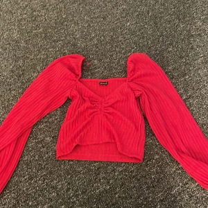 Röd ribbad topp med puffärmar - Trendig röd topp med puffiga långärmar och ribbad struktur. Toppen har en snygg rynkad detalj framtill som ger en unik look. Perfekt för att sticka ut i mängden!