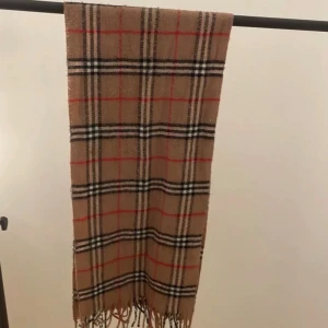 Burberry - Snygg rutig halsduk i beige med inslag av svart, vit och röd. Den har klassiska fransar i ändarna och är perfekt för att hålla dig varm under kyliga dagar.