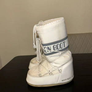 Vita Moon Boots- storlek 42-44 - Säljer ett par klassiska vita Moon Boots med snörning och grå detaljer.  De är endast använda under en vinter, har en liten defekt på baksidan (se bilder) men det är inget som syns, köpta för 2600 säljer för 1600🩵De är i storlek 42-44
