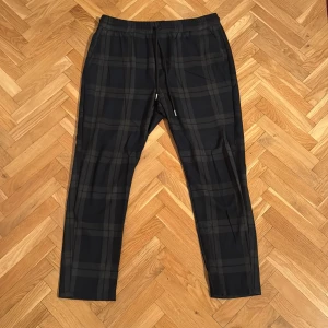 Flannel mönstrade kostymbyxor från Only & Sons - Använda kostymboxor med pyjamasbyx liknande design. Något midre i storlek. Snygga rutiga byxor från Only & Sons i svart och grått. Byxorna har en elastisk midja med snörning för extra komfort. Perfekta för en avslappnad stil med en modern touch.