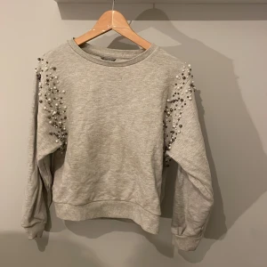 Grå sweatshirt med pärlor - Snygg grå sweatshirt med pärldetaljer längs ärmarna. Perfekt för att ge din outfit en unik touch. Tröjan har långa ärmar och en rund halsringning.