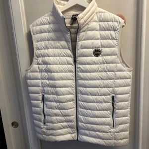 Vit dunväst från Colmar - Säljer en stilren vit dunväst från Colmar med dragkedja och två praktiska fickor framtill. Västen har en quiltad design och är perfekt för kyligare dagar. Den har en diskret logotyp på bröstet och passar bra över en hoodie eller tröja. Jackan har inga skador eller liknande men lite missfärgad på sidorna men inget man ser när den andvänds