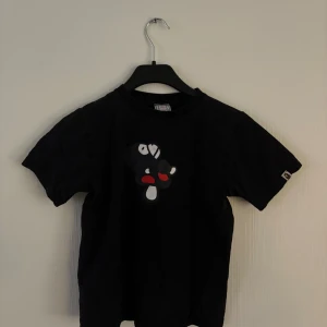 Svart t-shirt med tryck från A Bathing Ape - Cool svart t-shirt från A Bathing Ape med ett unikt tryck av en apa liggandes på en svamp i rött och vitt. Perfekt för dig som gillar streetwear och vill sticka ut med en stilren design. T-shirten har korta ärmar och en klassisk passform.
