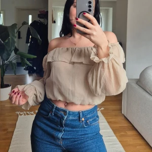Beige offshoulder blus från Bubbleroom - Säljer en söt beige offshoulder blus från Bubbleroom. Blusen har volangdetaljer och långa ärmar med elastiska ärmslut. Perfekt för en stilren och luftig look.