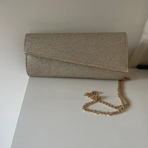 Glittrig balväska med guldkedja - Snygg glittrig balväska i silver med en elegant guldkedja. Perfekt för att ge din outfit en extra touch av glamour. Väskan har en stilren design med ett lock som stängs över framsidan.