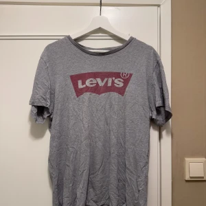 Grå t-shirt från Levi's - Säljer en grå t-shirt från Levi's med deras ikoniska logga i rött på bröstet. T-shirten har en rund halsringning och korta ärmar. 