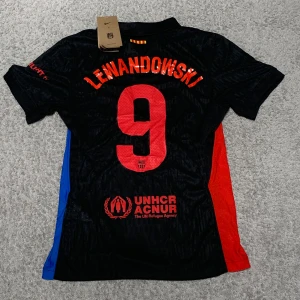 Svart fotbollströja med Lewandowski 9 - Snygg svart fotbollströja med Lewandowski 9 på ryggen. Tröjan har röda och blå detaljer på sidorna och är kortärmad. Den har även UNHCR-logga och Spotify-märke på framsidan.