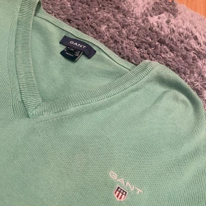 Grön v-ringad tröja från GANT - Snygg grön v-ringad tröja från GANT med broderad logga på bröstet. Tröjan har långa ärmar och ribbade muddar vid ärmslut och nederkant. Bara använd ett fåtal gånger och är i bra skick.