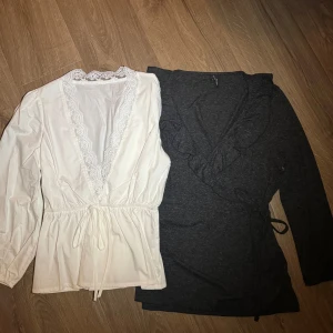 Två omlottblusar i vitt och grått - Säljer två snygga omlottblusar, en i vitt med spetsdetaljer och knytband i midjan, och en i mörkgrått med volangdetaljer. 