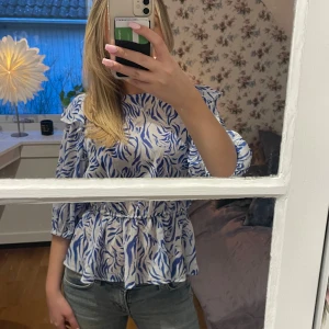 Blåmönstrad blus med volangdetaljer - Snygg blus från Kappahl (med prislapp kvar) med blått och vitt mönster. Blusen har volangdetaljer och en öppen rygg med knytband. Perfekt för en stilren look.