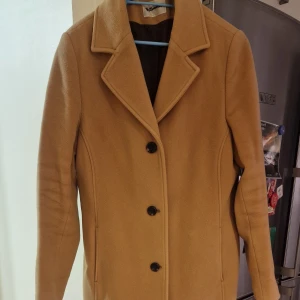 Beige kappa med knappar - Snygg beige kappa med klassisk design och svarta knappar. Perfekt för höst och vår. Den har långa ärmar och en stilren krage.