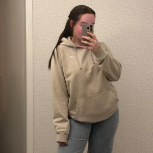 Beige hoodie från Gymshark - Säljer en snygg, beige hoodie från Gymshark med en broderad logga på bröstet. Tröjan har en klassisk känguruficka och justerbar huva. Perfekt för en avslappnad look.