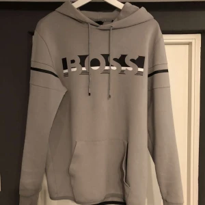 Grå hoodie från Boss - Snygg grå hoodie från Boss med svart och vit logga på bröstet. Jätte bra skick utöver snörrena som man ser