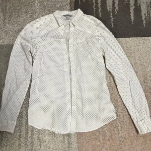 Vit prickig skjorta - Snygg vit skjorta med små svarta prickar. Skjortan har en klassisk krage och långa ärmar, perfekt för en stilren look. Passar bra till både jeans och kostymbyxor.