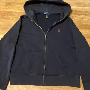 Navy Blå zip-hodie från Polo Ralph Lauren  - Säljer en stilren navy hoodie från Polo Ralph Lauren med dragkedja och röd broderad logga på bröstet. Tröjan är lite urtvättad men svårt att lägga märke till det. I sin helhet är tröjan i fint skick. Storlek 14-16 L i  barn storlek från kidsbrandstore. Säljer den för att jag har växt ur den