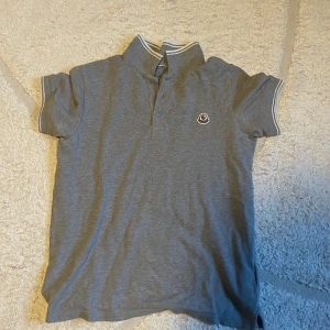 Grå pikétröja från Moncler - Säljer en stilren grå pikétröja från Moncler med korta ärmar och klassisk krage. Tröjan har vita detaljer på kragen och ärmsluten samt Monclers logotyp på bröstet. Perfekt för en avslappnad men ändå snygg look.