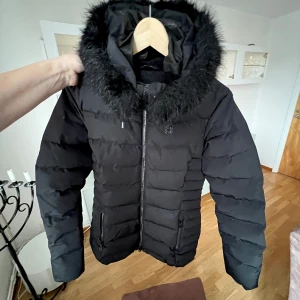Svart dunjacka med pälskrage - Snygg svart dunjacka med dragkedja och pälskrage. Lång svart dunjacka i bra skick. Pälsen på luvan är borttagnings bar. Orginalpris 3000 kr