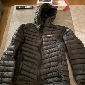 Svart dunjacka från Peak Performance - Säljer en svart dunjacka från Peak Performance. Jackan har en quiltad design med dragkedja och huva. Perfekt för kyliga dagar och håller dig varm och bekväm. En stilren och funktionell jacka för alla tillfällen.