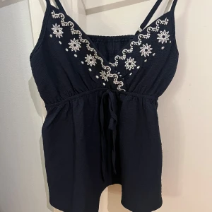 Svart ärmlös topp med broderi - Snygg svart ärmlös topp med vita blombroderier och knytband framtill. 