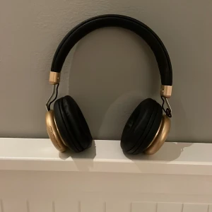 Svarta och guldiga hörlurar - Snygga hörlurar i svart och guld med vadderade öronkuddar för extra komfort. De har knappar för volymkontroll och uppspelning på sidan. Perfekta för musikälskare som vill ha stil och funktionalitet.