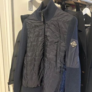 Mörkblå cardigan från J.Lindeberg - Snygg mörkblå cardigan från J.Lindeberg med quiltad framsida och stickade ärmar. Jackan har en dragkedja framtill och en hög krage. Perfekt för kyligare dagar. 