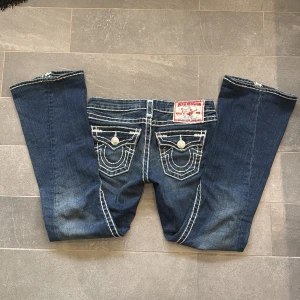 Mörkblå jeans från True Religion - Snygga mörkblå jeans från True Religion med kontrasterande vita sömmar och klassiska femficksdesign. Jeansen har en låg midja och är flared. Perfekt för en avslappnad stil. Märkets logga syns tydligt på bakfickan och etiketten. Innerben:76 Midjemått: 37. Inga bilder på.