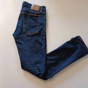 Mörkblå jeans från Nudie Jeans - Snygga mörkblå jeans från Nudie Jeans med klassisk femficksdesign och orange sömmar. Jeansen har en normal passform och är tillverkade i slitstarkt denim. Perfekta för en stilren look. Lappen på modellen är avklippt. Hör gärna av dig vid minsta fundering!