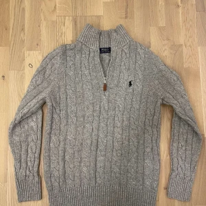 Grå stickad tröja från Ralph Lauren - Säljer en stilren grå stickad tröja från Ralph Lauren med kabelstickat mönster och dragkedja vid halsen. Tröjan har långa ärmar och ribbade muddar. Perfekt för kyliga dagar! Passar dig som har storlek S-M Det går också att diskutera om priset