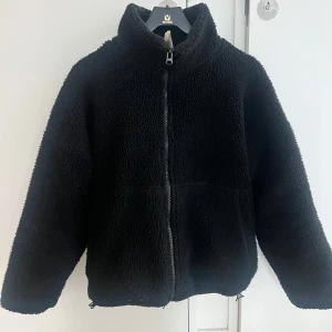 Svart teddyjacka med dragkedja - Mysig svart teddyjacka med dragkedja framtill. Jackan har en hög krage och långa ärmar, perfekt för kyliga dagar. Den är gjord i ett mjukt och fluffigt material som håller dig varm och bekväm.